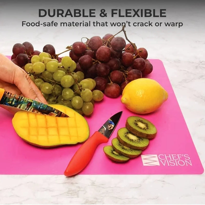 A hand slices a mango on a pink Chef’s Vision Slice Bright Flexible Cutting Mat, with grapes, lemon, and kiwis nearby. Two knives and the Chef’s Vision logo are seen. Text highlights the durability of these BPA-free mats.