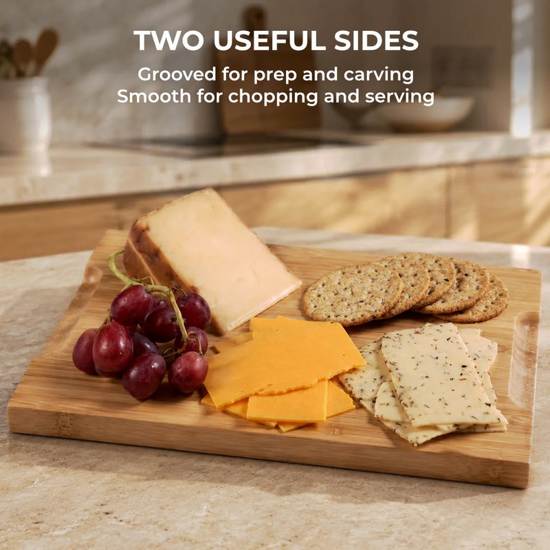 On a kitchen counter, a SliceBright Moso Bamboo Cutting Board by Chef's Vision displays grapes, two types of sliced cheese, crackers, and a cheese wedge within its juice grooves. Text above highlights the board’s two versatile sides and knife-friendly design.