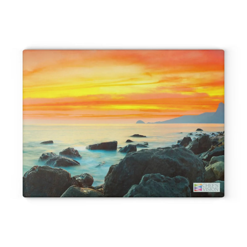 Vivid orange-yellow sunset over rocky shore and calm sea with distant mountains—a stunning scenic design on the SliceBright Landscape Glass Cutting Board by Chef's Vision, featuring their logo in the bottom right; ideal for your kitchen counter.
