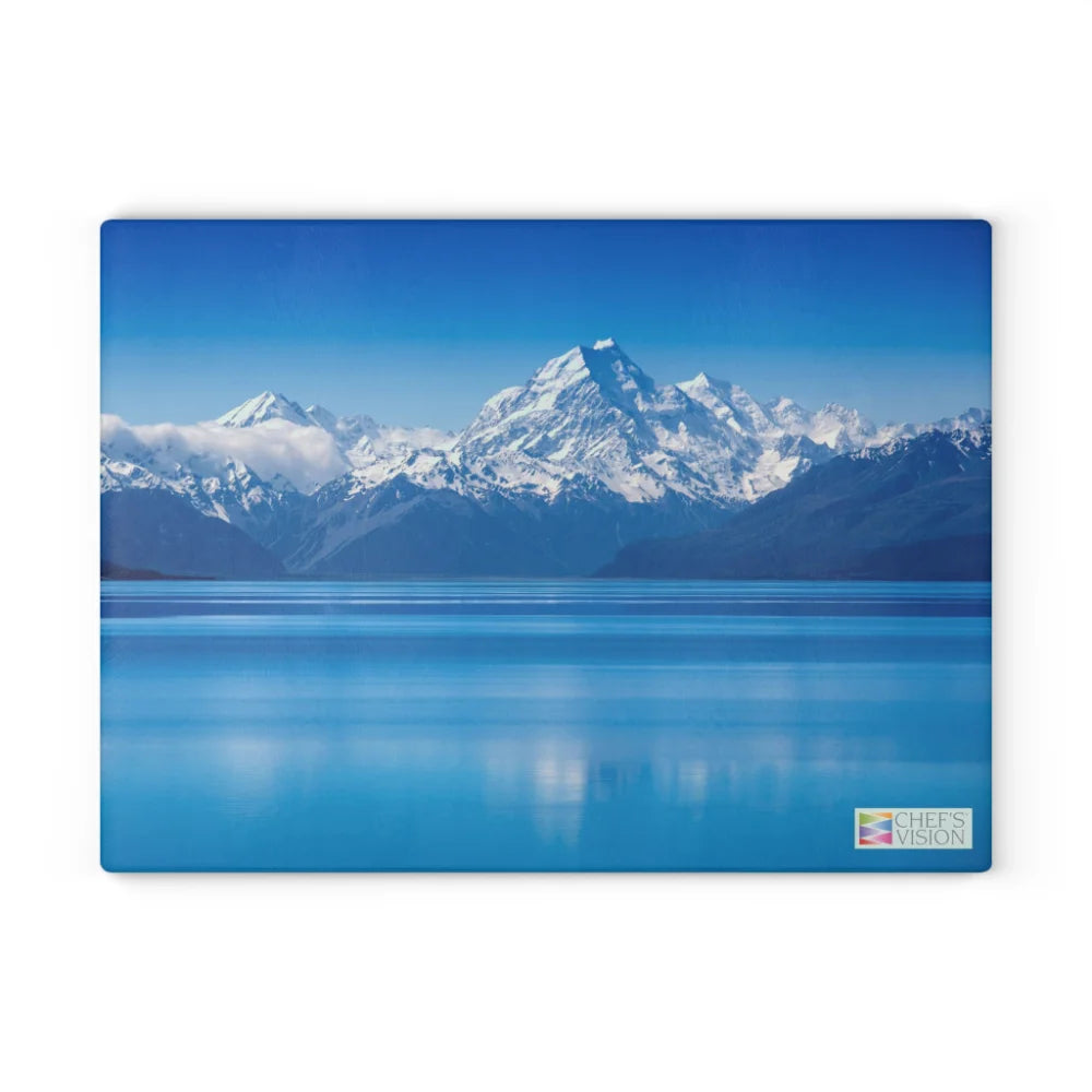 Snowy mountains mirrored in a calm blue lake beneath a clear sky—this scenic image graces the SliceBright Landscape Glass Cutting Board by Chef’s Vision. A small Chef’s Vision logo appears in the lower right corner, adding elegance to any kitchen.