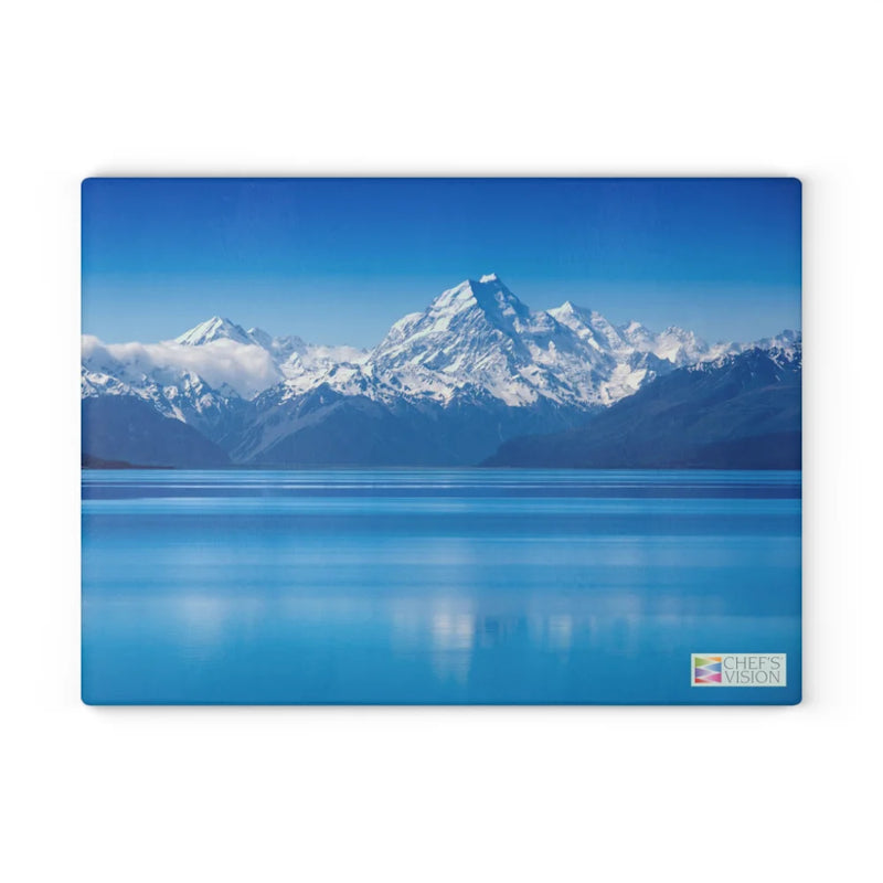 Snowy mountains mirrored in a calm blue lake beneath a clear sky—this scenic image graces the SliceBright Landscape Glass Cutting Board by Chef’s Vision. A small Chef’s Vision logo appears in the lower right corner, adding elegance to any kitchen.