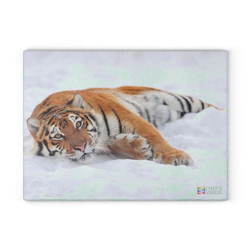A tiger rests in the snow, gazing at the camera—an image that highlights the majesty of wildlife on the Chef's Vision SliceBright Wildlife Glass Cutting Board.