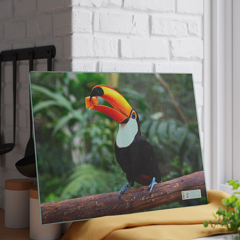 A toucan perched on a branch, with a Chef's Vision SliceBright Wildlife Glass Cutting Board displayed nearby.