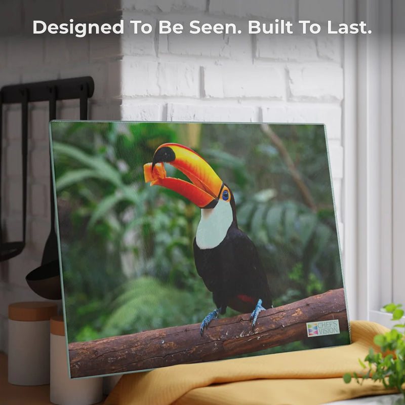 The SliceBright Wildlife Glass Cutting Board by Chef's Vision, featuring a vibrant toucan with an orange beak, is showcased on a wooden surface before a white brick wall. Text above: “Designed To Be Seen. Built To Last.” Ideal for wildlife art lovers.