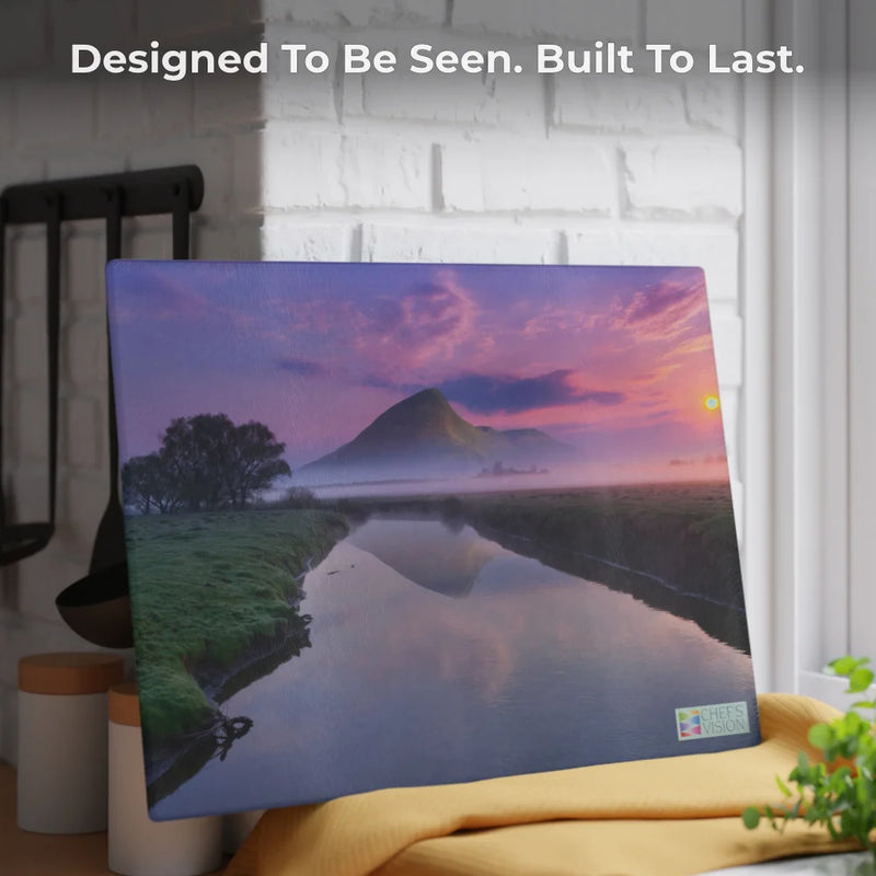 The SliceBright Landscape Glass Cutting Board by Chef's Vision features a scenic sunset with river, trees, and mountain. Displayed on a kitchen counter, it reads: “Designed To Be Seen. Built To Last.” Perfect match for canvas prints.